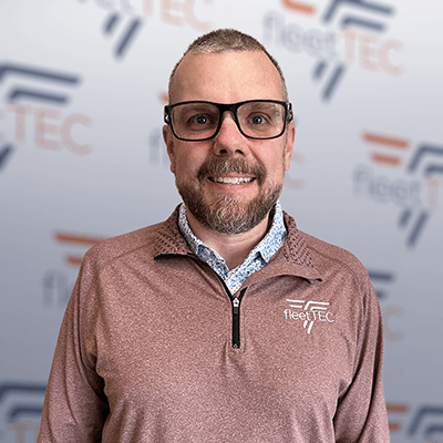 Man in glasses smiling, wearing a maroon FleetTEC shirt, standing before a FleetTEC logo backdrop.