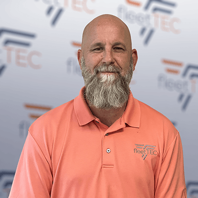 Man in a coral shirt with Fleet TEC logo, standing against a branded wall, smiling gently with a gray beard.