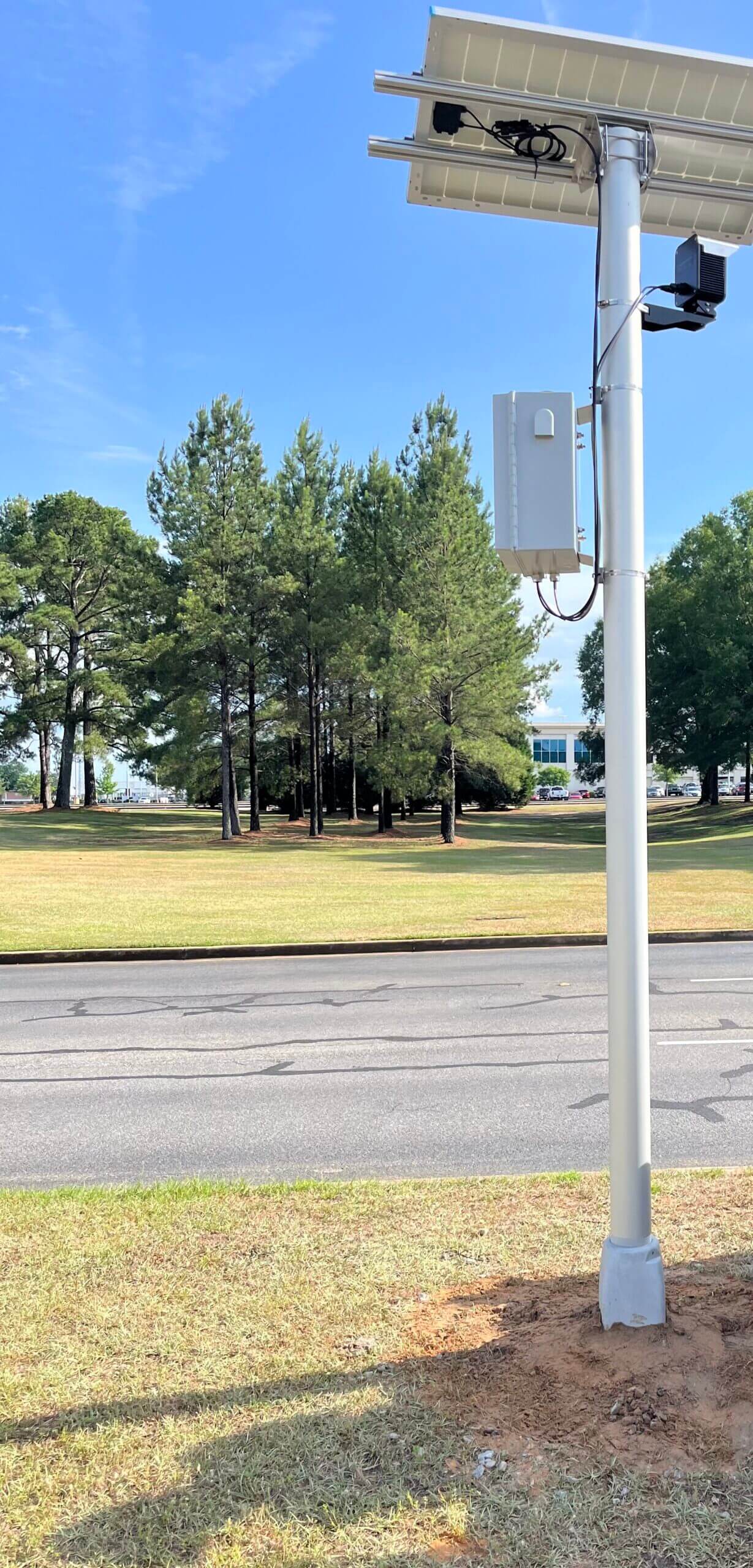 Solar-powered technology pole near a road, with trees and distant buildings in the background.